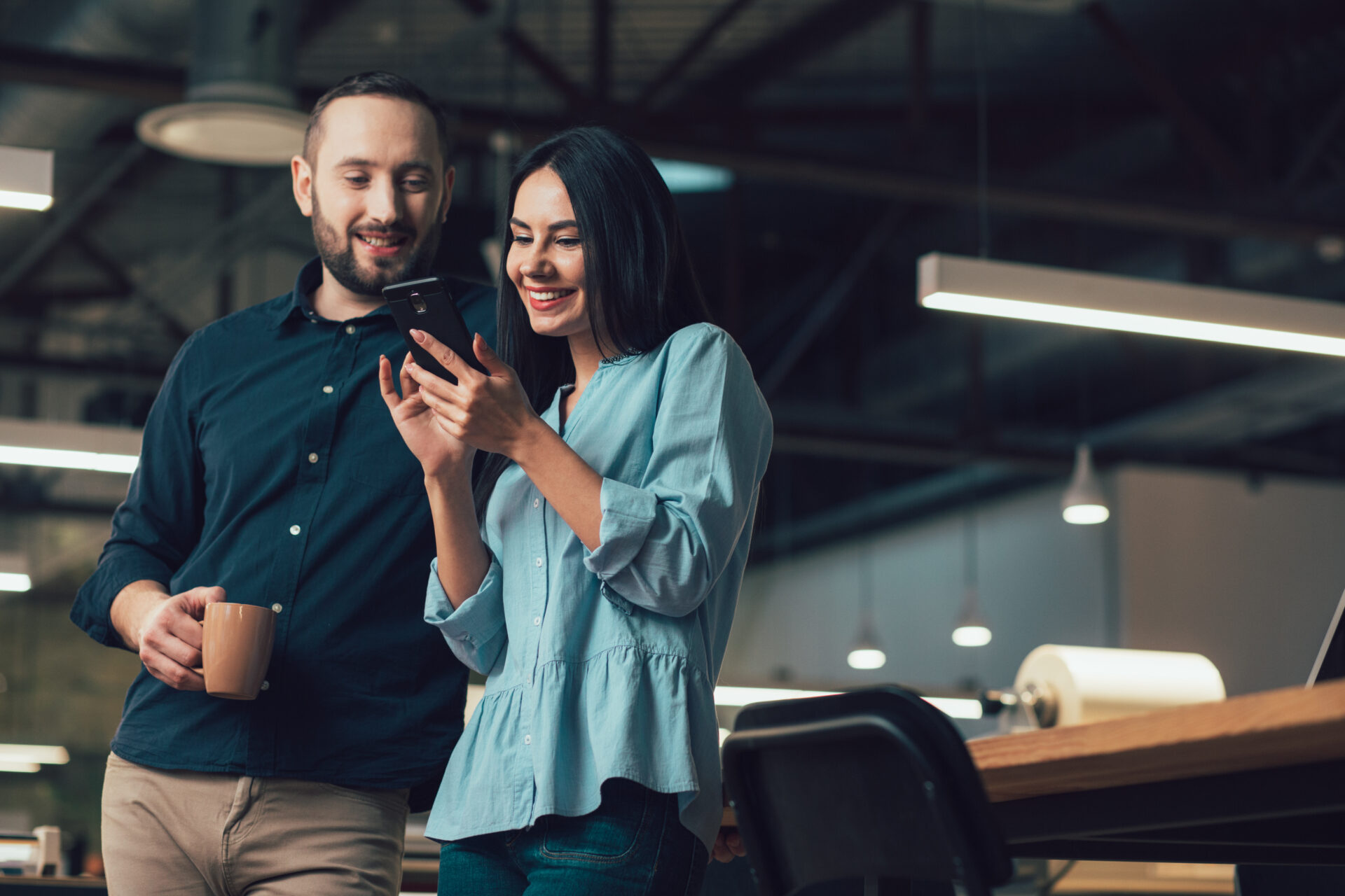 Happy lady using a modern smartphone and a cheerful man standing by her side with a mug and looking at her gadget